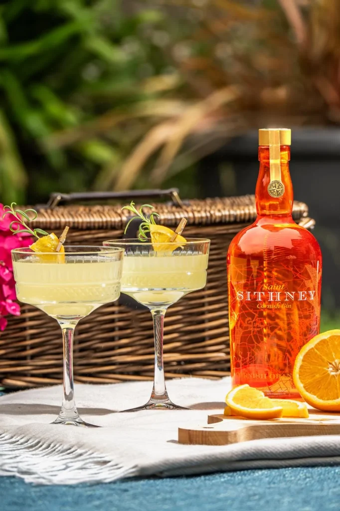 Sithney Bottles Of Gin And Oranges On A Table With A Basket Of Flowers